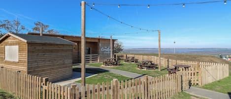 A wooden cabin with a sign reading 'The Creative Studio,' surrounded by picnic tables and a wooden fence, with a clear view of the water in the background.