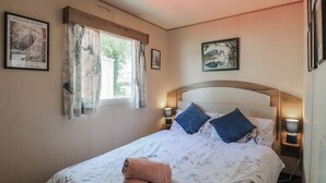 A bedroom with a bed, two wall-mounted lamps, a window with curtains, and two framed pictures on the wall.