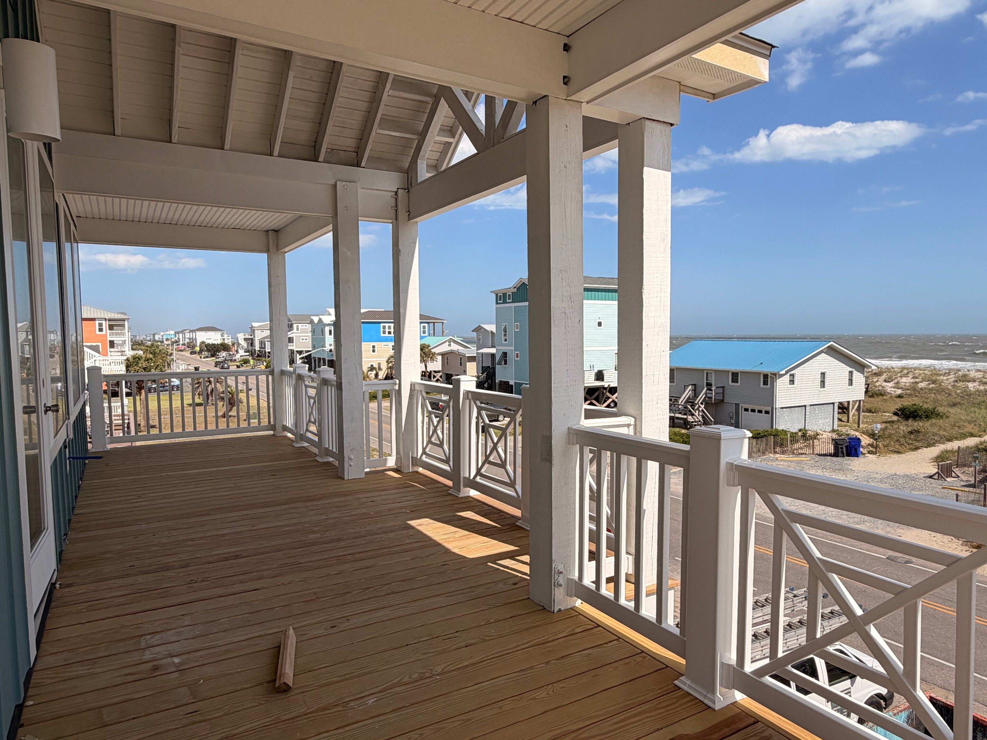 A wooden deck with white railings, a clear view of the beach, and houses in the background.