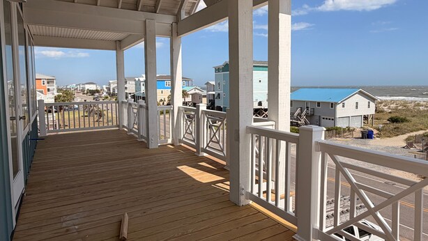 Eine Holzterrasse mit weißen Geländern, freier Blick auf den Strand und Häuser im Hintergrund.