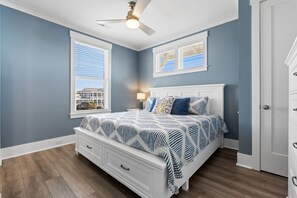 Ein Schlafzimmer mit einem Bett, zwei Fenstern mit Jalousien, einem Deckenventilator, einem Nachttisch mit Lampe und einer Kommode.