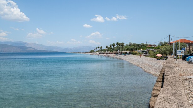 Escena costera de playa con agua cristalina, playa de guijarros y montañas a lo lejos.