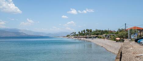 A coastal beach scene with clear water, a pebble beach, and distant mountains.