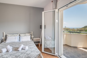 Un dormitorio con cama, mesita de noche, cajón, puerta de cristal que da a un balcón y vistas al paisaje.