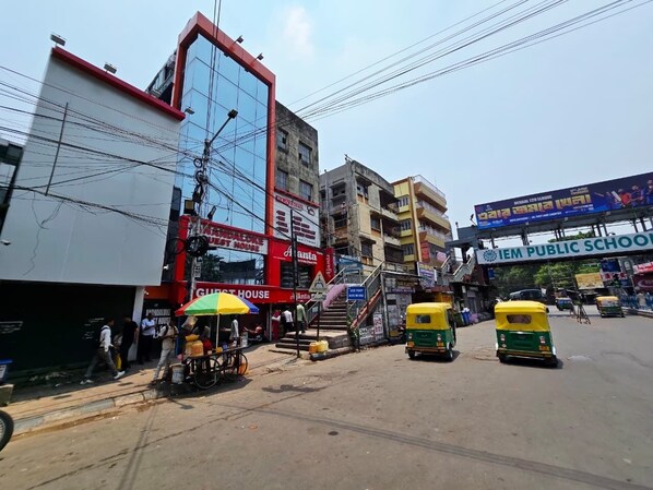 Una escena callejera con tuk-tuks, un edificio de fachada roja y blanca, y un letrero de escuela pública.