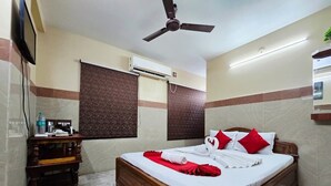 A hotel room with a bed, a ceiling fan, an air conditioning unit, a wooden nightstand, a black bag, and a patterned rug. - Anandaloke Guest House (Kolkata)