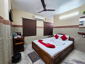 Una habitación de hotel con cama, ventilador de techo, equipo de aire acondicionado, cómoda de madera y una pequeña alfombra.