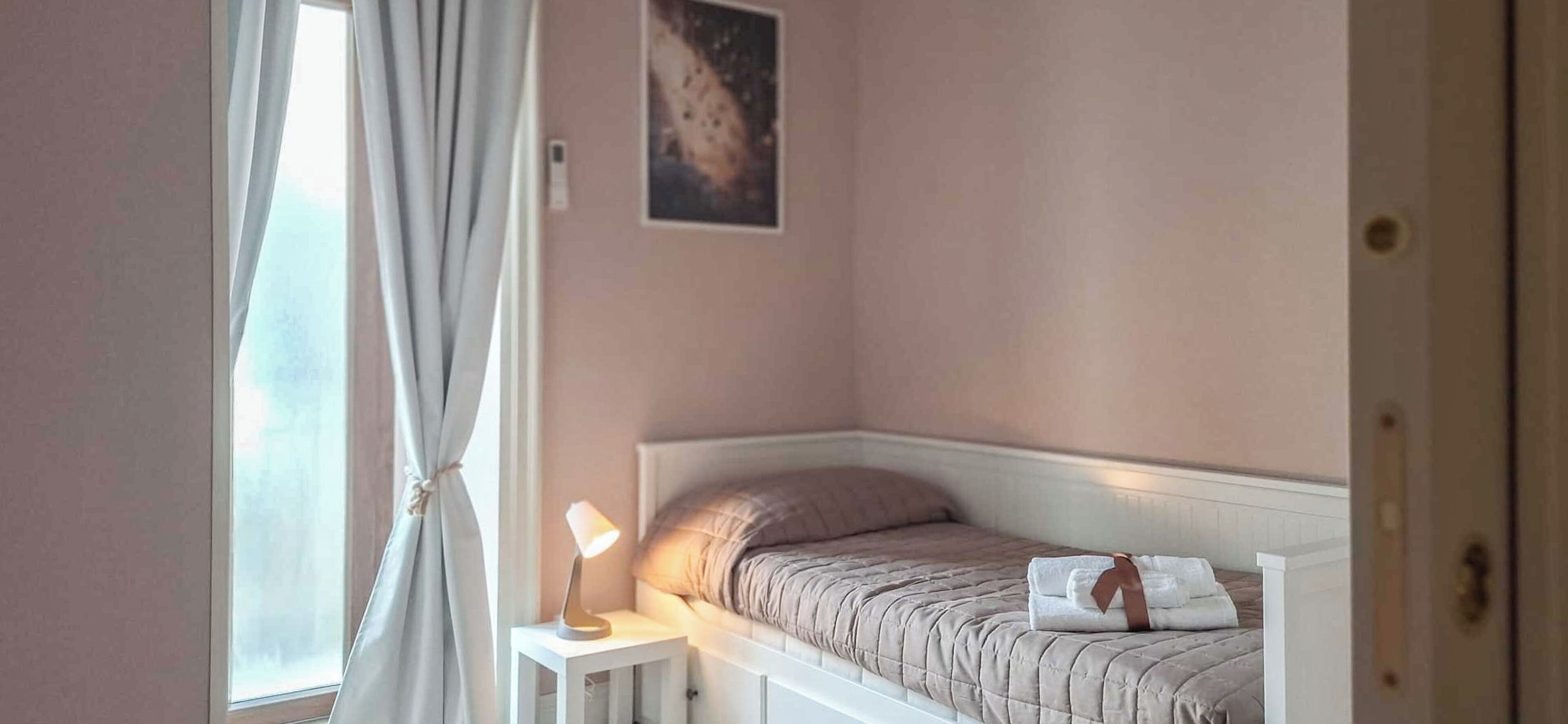 A bedroom with a bed, a bedside lamp, a window with curtains, and a wall-mounted picture.