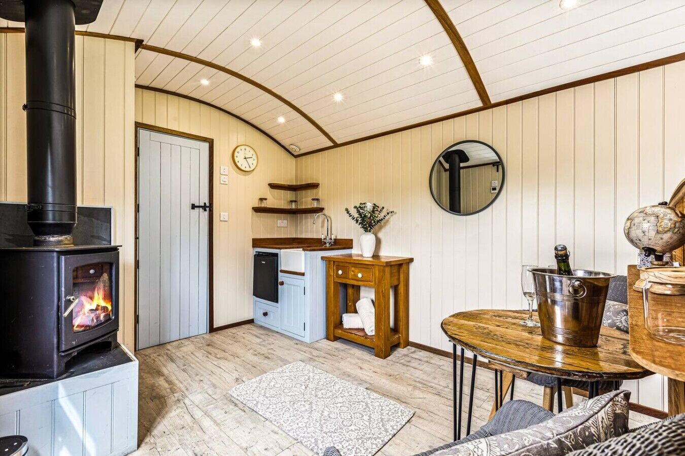 Een gezellige kamer met een houtkachel, een eettafel, een klein keukenhoekje en een ronde spiegel.
