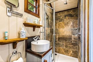 Casa de banho com uma pia branca, cabine de chuveiro em vidro e uma prateleira de madeira com artigos de higiene pessoal.