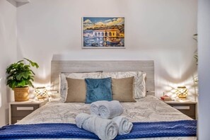 A neatly made bed with rolled towels, a headboard, and a painting of a bridge over water. - Valletta Marina View - Seafront Apartment (Pietà)