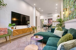 A modern living room with a green sofa, a wooden console table, a flat-screen TV, and a dining area in the background. - Casa Botanica, charming Maltese townhouse (Luqa)