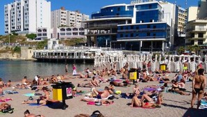 En strand med folk som solar, ett vattendrag och byggnader i bakgrunden.