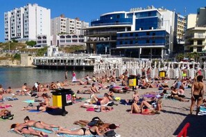 Ein Strand mit badenden Personen, einer Wasserfläche und Gebäuden im Hintergrund.
