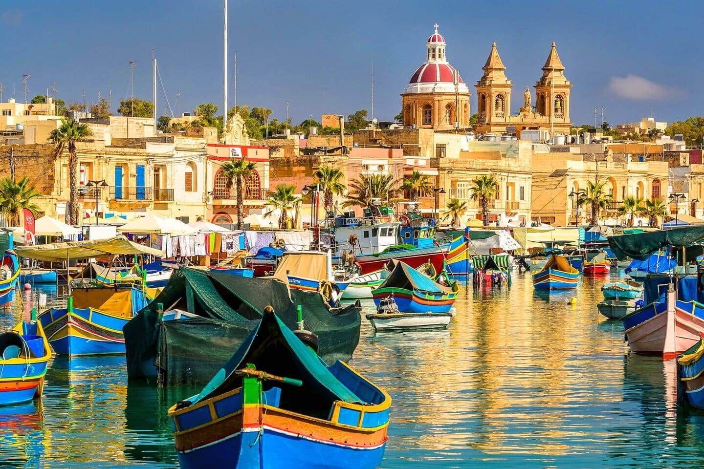A marina with numerous colorful boats docked, a prominent church with a red dome, and a coastal townscape in the background.