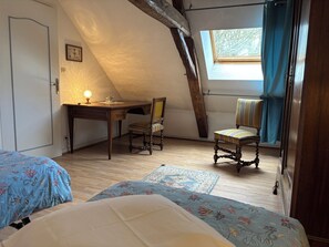 Une chambre avec un plafond en pente, un bureau avec une lampe, deux chaises, un lit avec une literie à motifs floraux et une fenêtre avec des rideaux.