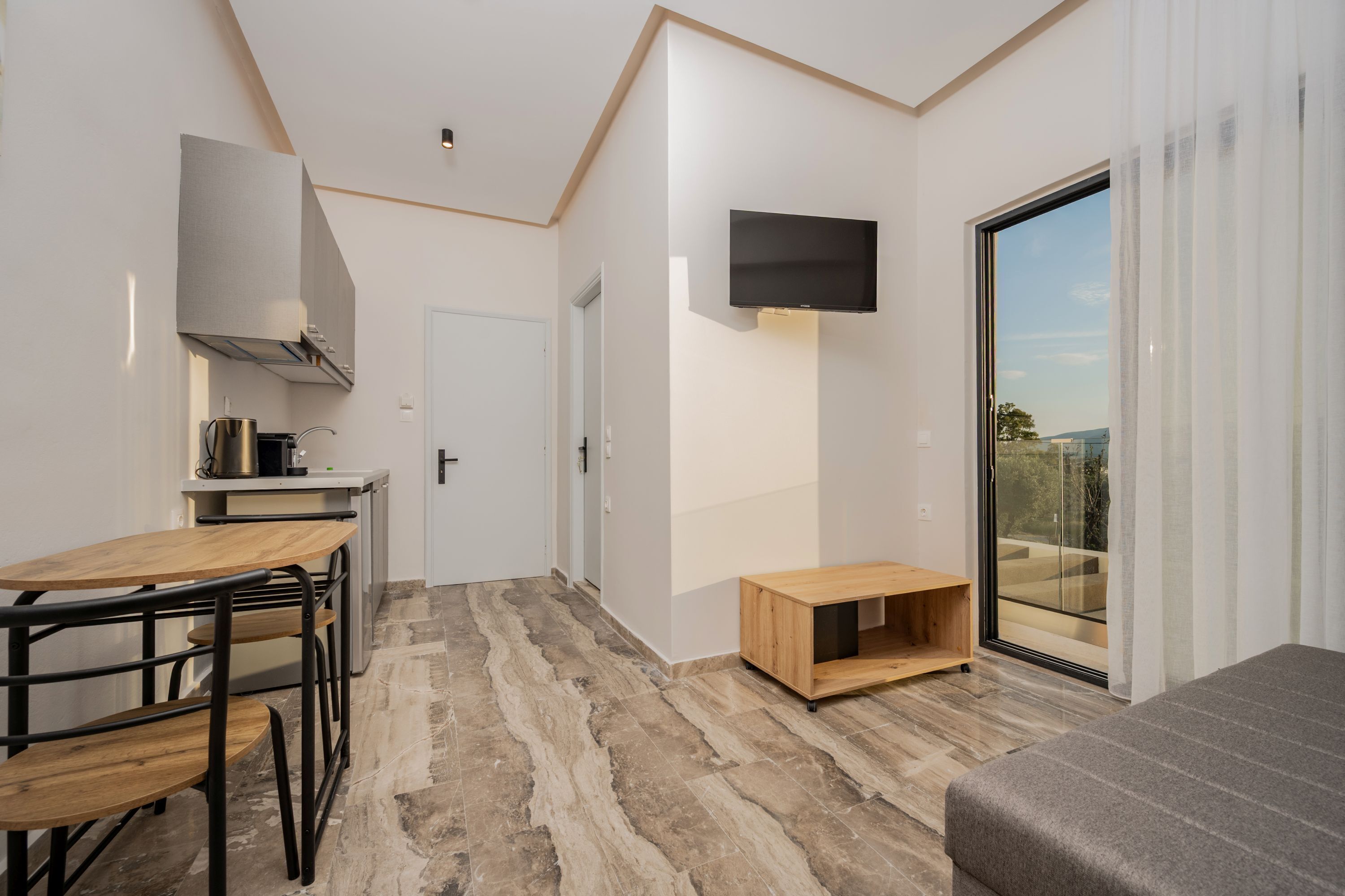 A modern room with a kitchen area, a dining table, a TV mounted on the wall, and a sliding glass door leading to an outdoor balcony.