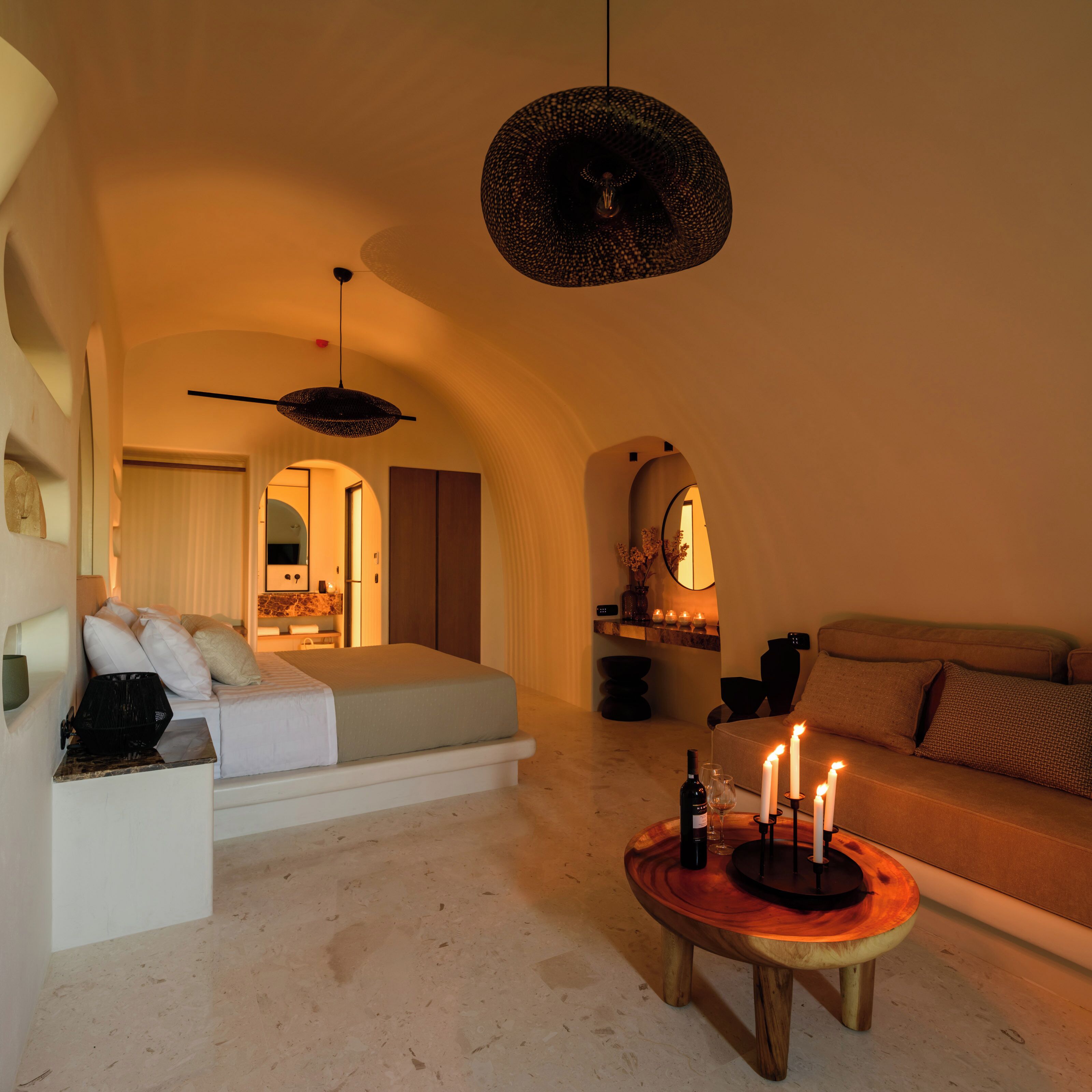 A cozy hotel room with a bed, a sofa, a round wooden coffee table with candles, and a hanging light fixture.
