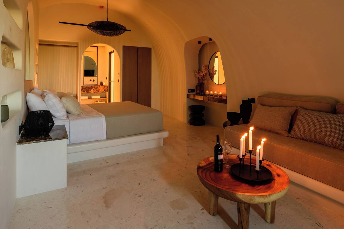 Une chambre d’hôtel confortable avec un lit, un canapé, une petite table basse ronde en bois ornée de bougies et un luminaire suspendu.
