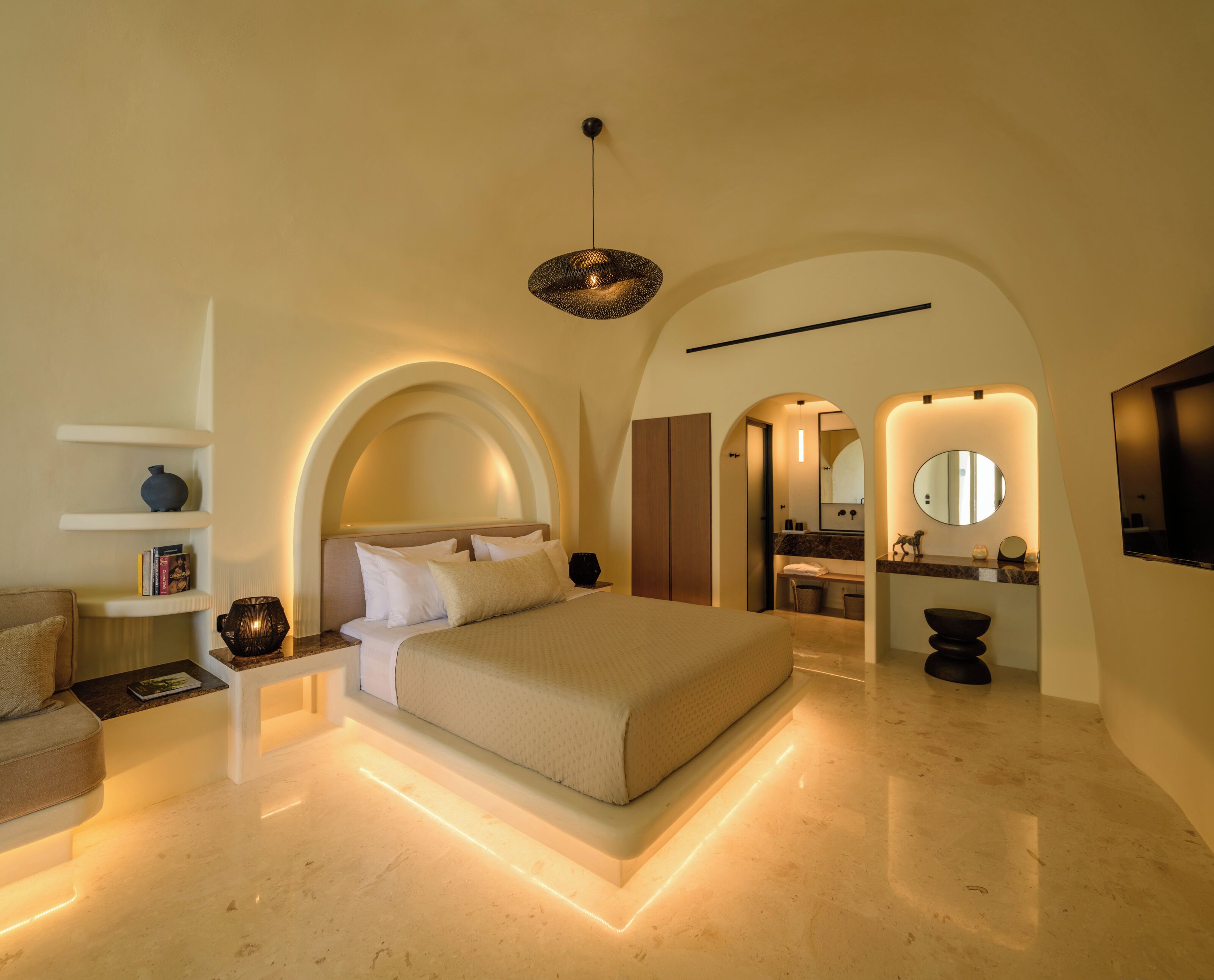 A modern bedroom with a large bed, bedside tables, a reading lamp, and a bathroom with a mirror and sink.