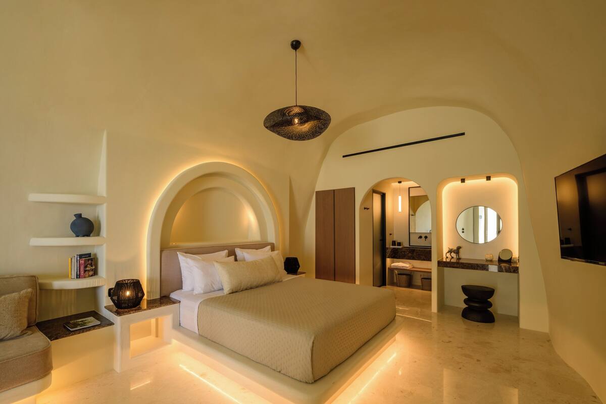 Une chambre à coucher moderne avec un grand lit, des tables de chevet, une lampe de lecture et une salle de bain équipée d’un miroir et d’un lavabo.