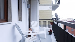Ein Balkon mit weißen Plastikstühlen und einem Tisch, einem Blumentopf mit roten Blüten sowie einer Satellitenschüssel am Gebäude.