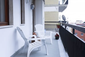 Un balcone con sedie e tavolo in plastica bianca, una pianta in vaso con fiori rossi e un'antenna parabolica sull'edificio.