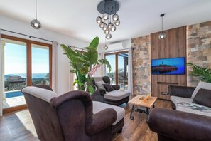 Ein modernes Wohnzimmer mit großem Fernseher, bequemen Sitzmöglichkeiten, einer Pflanze und Blick ins Freie.