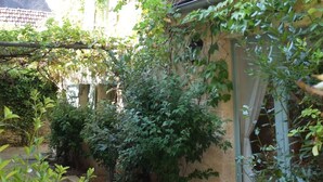 Een landelijk gebouw met een stenen gevel, een raam met gordijn, en een tuin met verschillende planten en een waterslang.