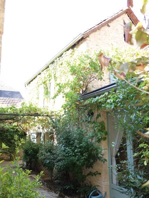 Um edifício rústico com fachada de pedra, uma janela com cortina e um jardim com diversas plantas e uma mangueira.