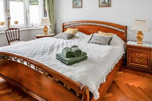 Una cama bien tendida con colcha, almohadas y toallas enrolladas, una mesita de noche de madera con una lámpara, una silla y una ventana con cortinas.