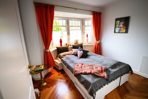 Ein Schlafzimmer mit Bett, einem Fenster mit Vorhängen, einem Nachttisch und einem an der Wand hängenden Bild.