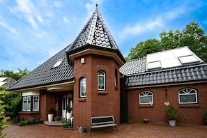 Une maison en briques avec un toit conique, une véranda à l’avant et un petit jardin.