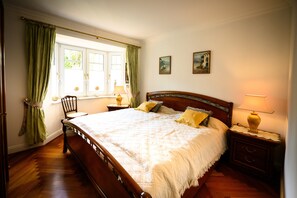 A bedroom with a wooden bed, green curtains, two paintings on the wall, and a nightstand with a lamp.