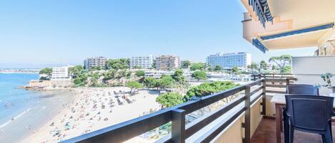 A balcony with a view of a beach, a dining area with chairs, and a tiled floor.
