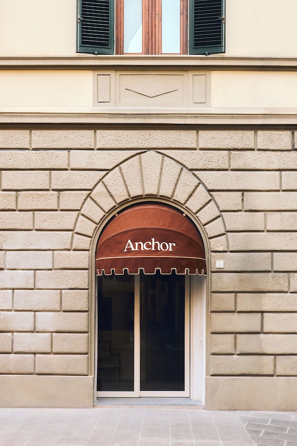 A building facade with a brick wall, a brown awning with the word 'Anchor' above a glass door entrance.