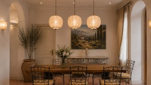 Une salle à manger avec une table en bois, quatre chaises, un vase de fleurs, un buste posé sur la cheminée et un tableau représentant un paysage accroché au mur.