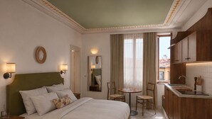 Une chambre d’hôtel comprenant un lit, des tables de chevet, une petite table ronde avec des chaises, un miroir, une fenêtre avec des rideaux et un coin cuisine.