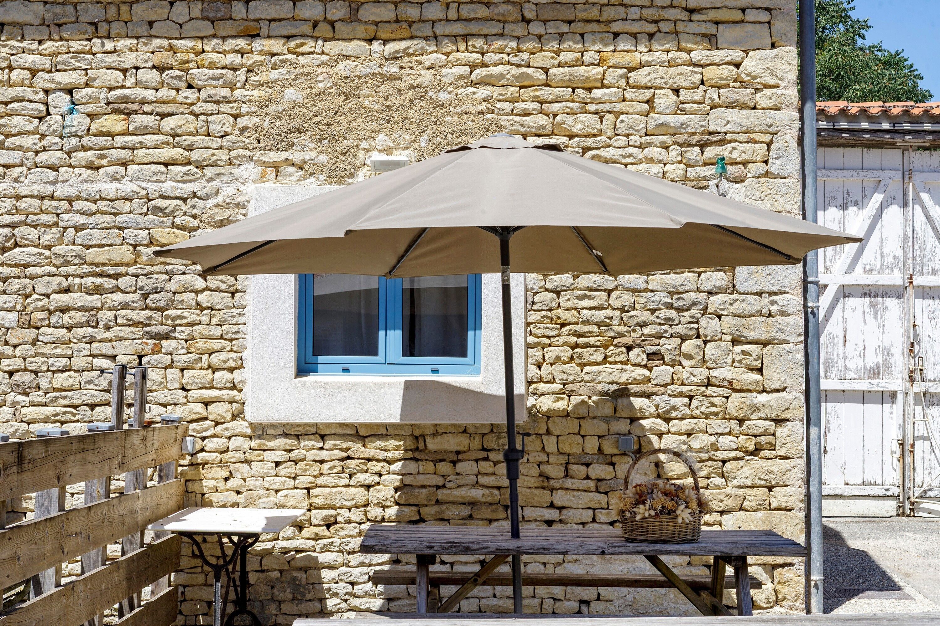 Un edificio de piedra con una mesa y banco de madera, una ventana con contraventanas azules y un paraguas grande.