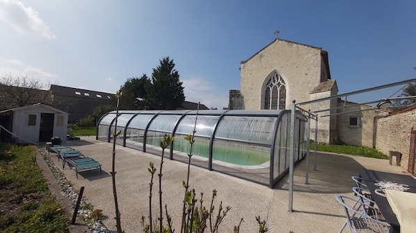 A greenhouse structure with a transparent roof, a stone building with a pointed arch window, and a small shed in the background.