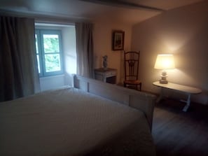 A bedroom with a bed, a window with curtains, a chair, a small table with a lamp, and a picture on the wall.