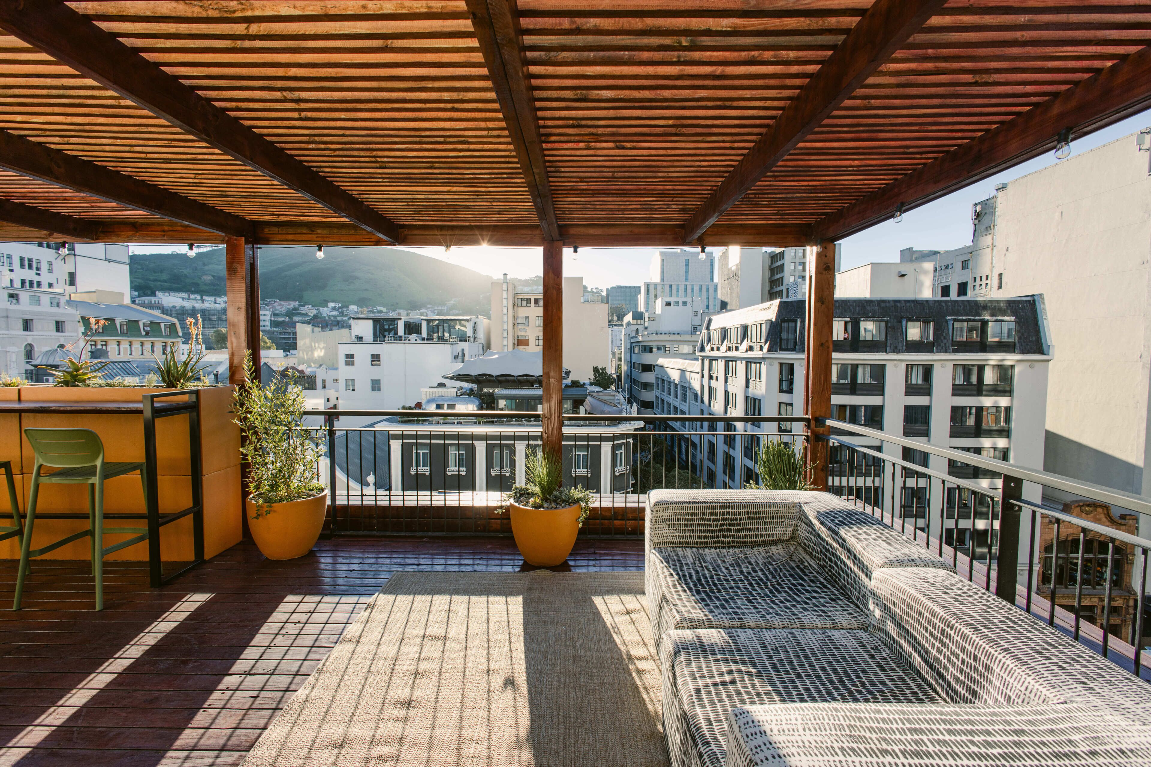 A balcony with a wooden pergola, two potted plants, a wicker sofa, and a view of the cityscape.