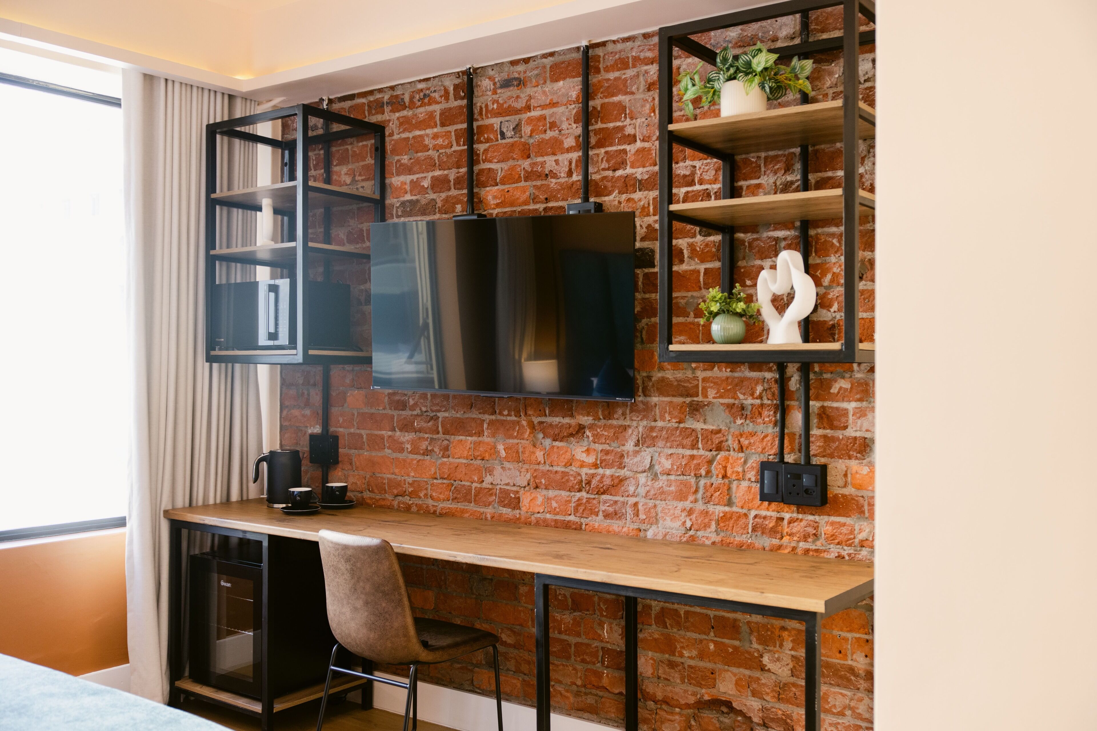 A modern room with a brick wall, a wooden desk, a black chair, a television mounted on the wall, and a shelf with decorative items.