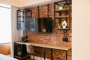 Una habitación moderna con pared de ladrillo, un escritorio de madera, una silla negra, un televisor montado en la pared y un estante con objetos decorativos.