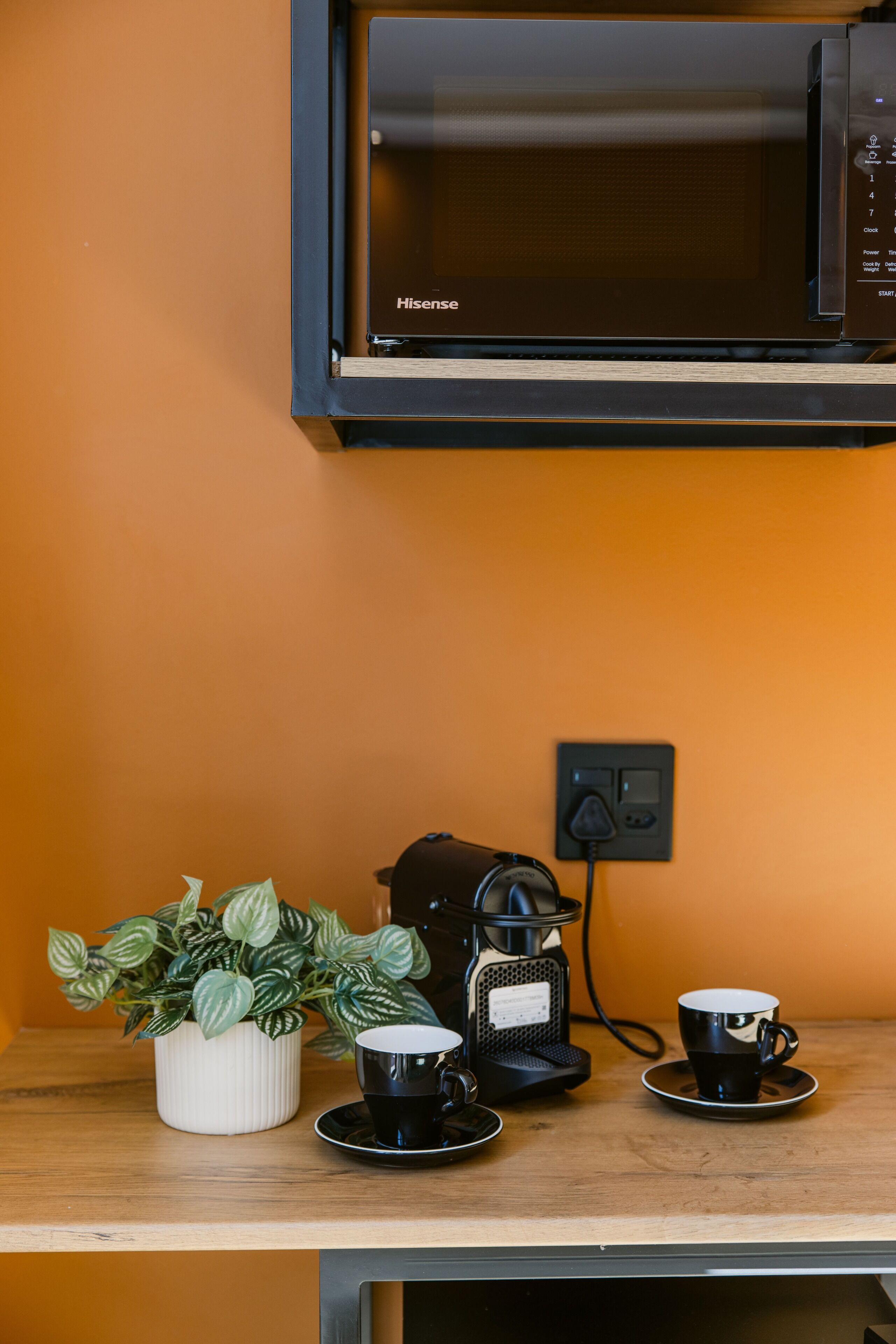 Un coin cuisine équipé d’un four à micro-ondes, d’une machine à café et de deux tasses posées sur une table en bois.