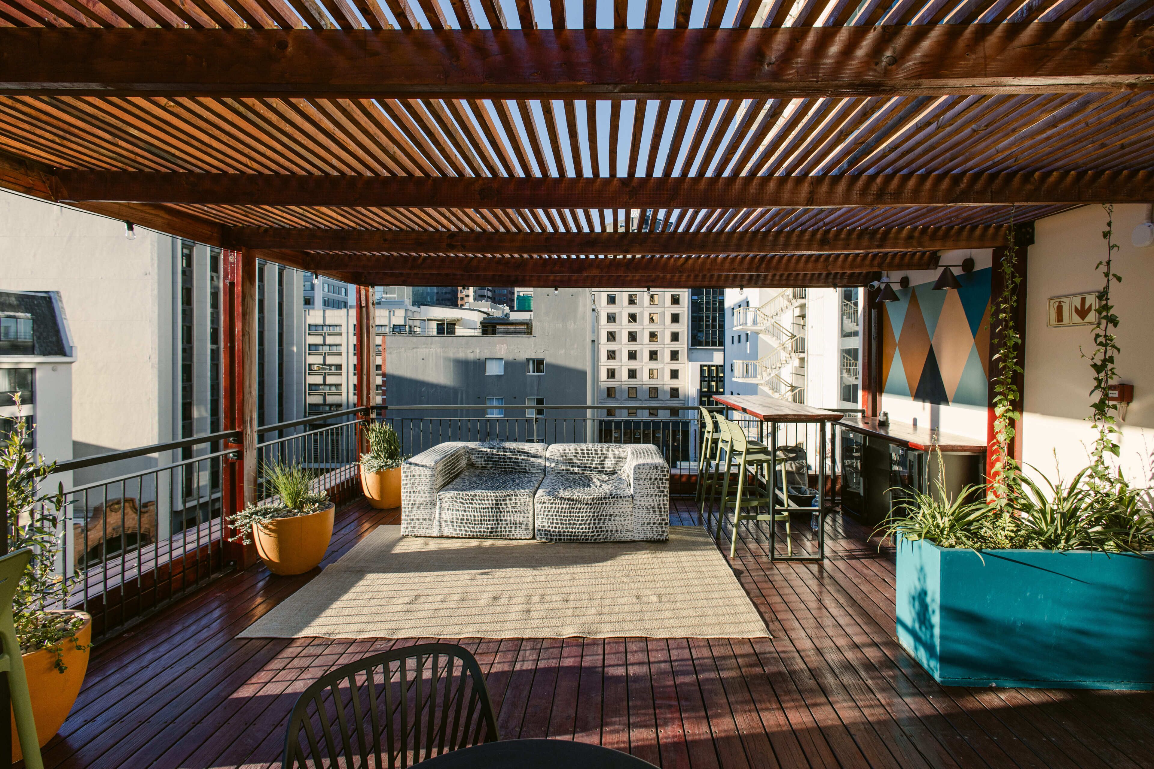 Un balcone con una pergola in legno, posti a sedere all'aperto, piante in vaso e vista sugli edifici della città.