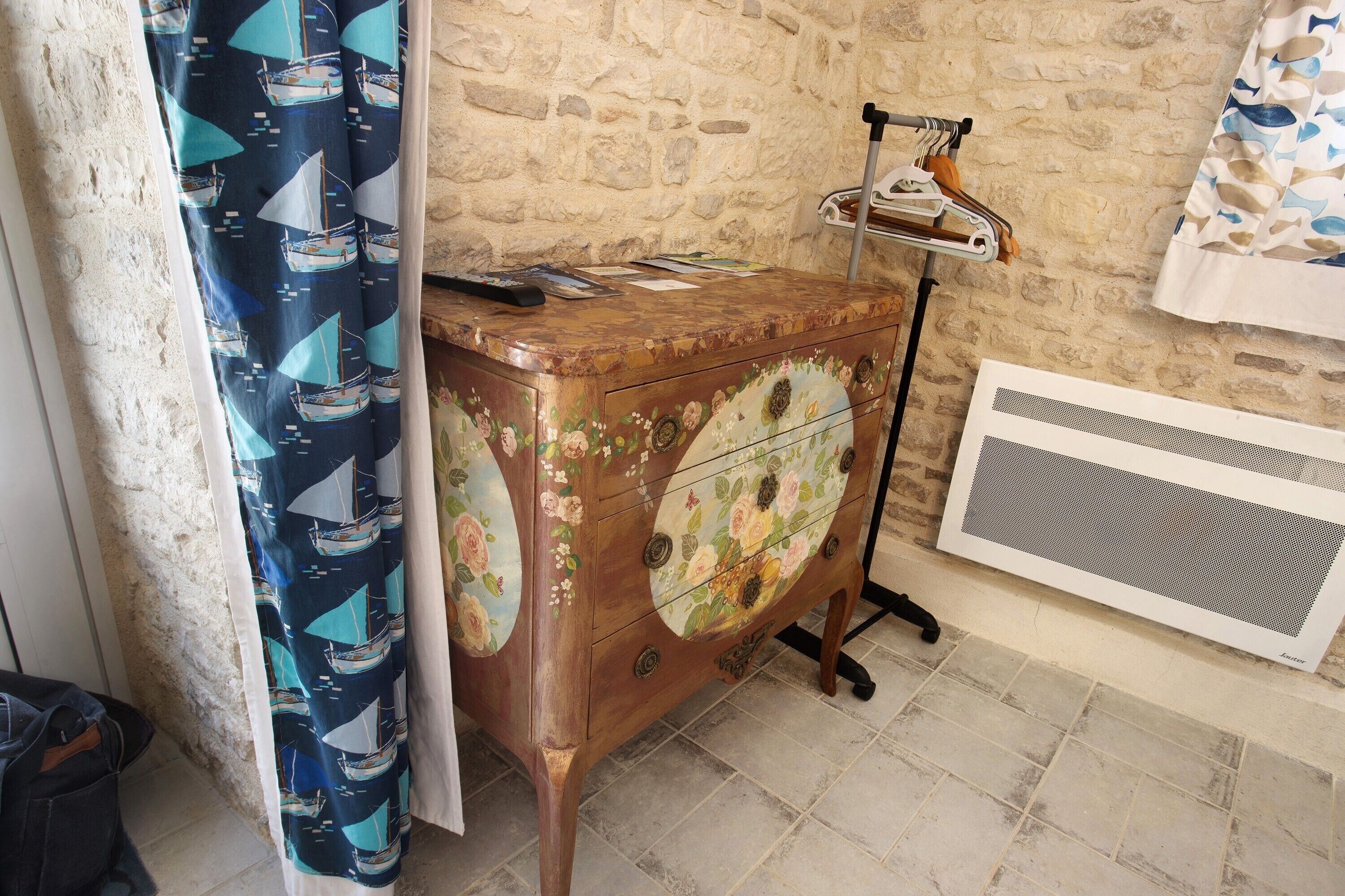 Eine rustikale Steinwand, ein Holzschrank mit floralen und Segelbootmotiven, eine Heizung und ein Vorhang mit Bootsdesigns.