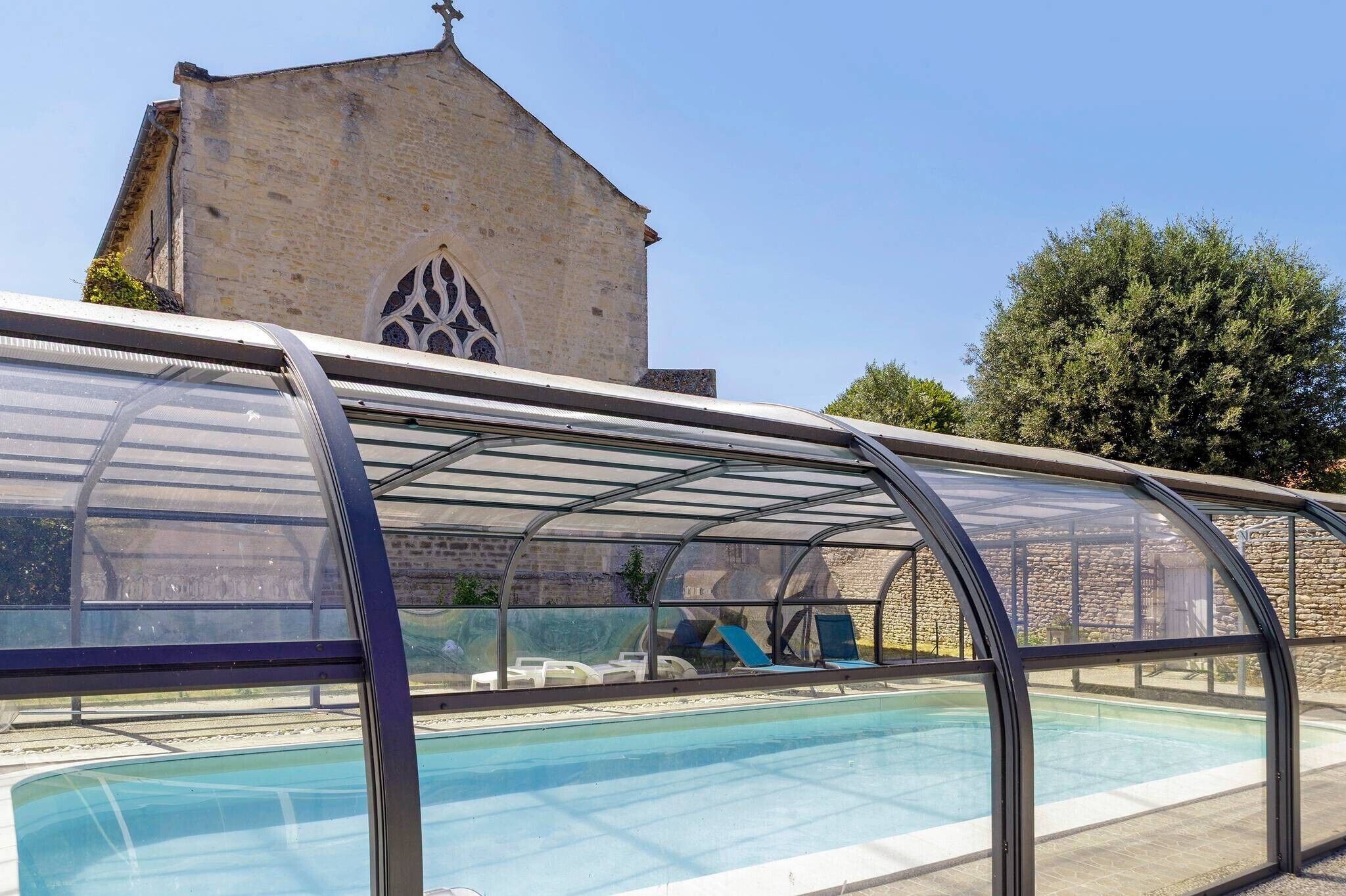 Uma piscina com cobertura de vidro, um edifício de pedra com uma cruz no topo e árvores ao fundo.