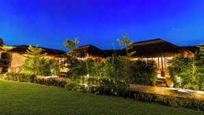 A nighttime view of a resort with thatched-roof buildings, well-lit pathways, and lush greenery. - ELIVAAS Kimaya Suite With Game Zone (Gurugram)