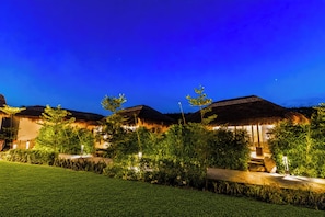 A nighttime view of a resort with thatched-roof buildings, well-lit pathways, and lush greenery. - ELIVAAS Kimaya Suite With Game Zone (Gurugram)